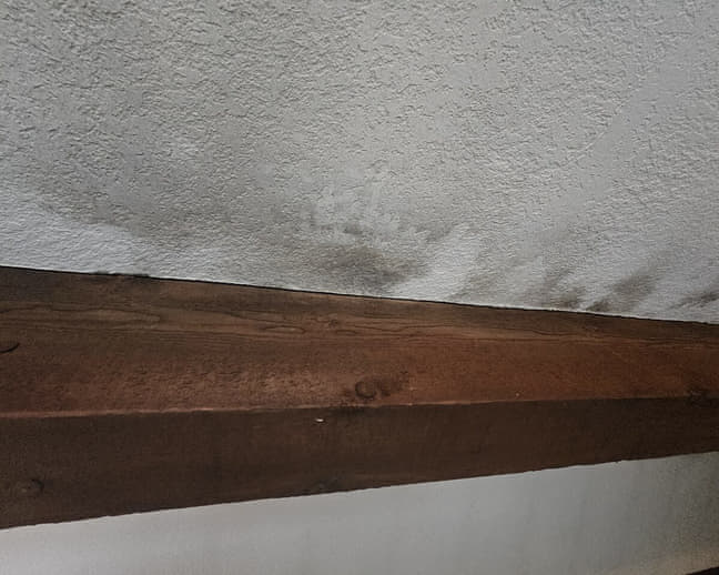 Water stain on a textured ceiling beside a dark wood beam from a roof leak in Rockford IL