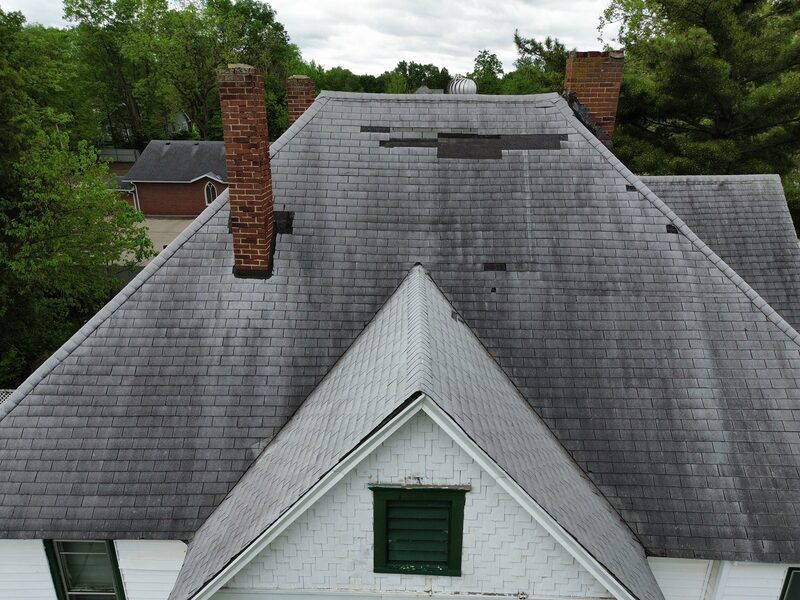 Aerial view of an aged asphalt roof in Decatur, Illinois with multiple scattered missing shingles across two main slopes, weathered gray color, two brick chimneys and a front gable dormer above white wood siding