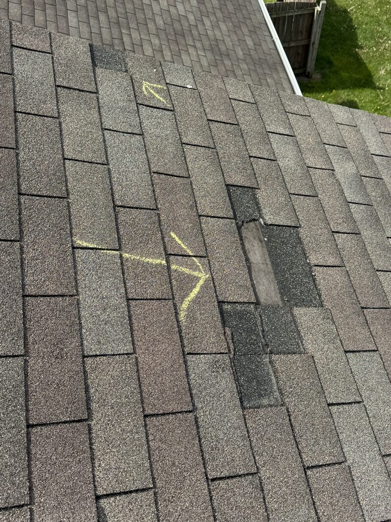 Asphalt shingle slope with two missing shingles after a wind event, exposed underlayment showing through, yellow chalk arrows marking each loss for the adjuster