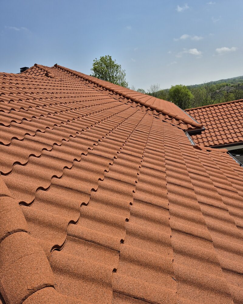 Broad slope of terracotta stone-coated metal tile rising to a hip ridge, a second roof plane and treed hillside beyond, Brookfield WI