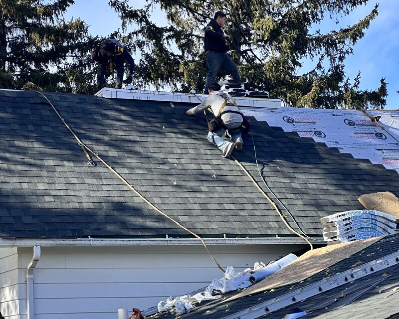 Roofing crew installing new charcoal asphalt shingles during a roof replacement on a Beloit WI home, with synthetic underlayment still exposed on the upper slope