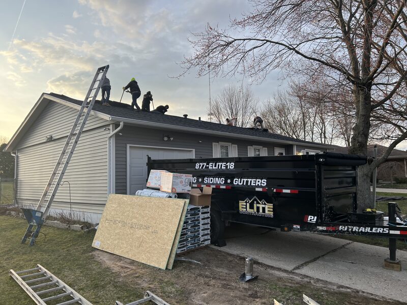 Roofing crew tearing off old shingles on a grey ranch in Loves Park IL, with IKO Cambridge shingle bundles and an Elite Contracting dump trailer staged on the driveway