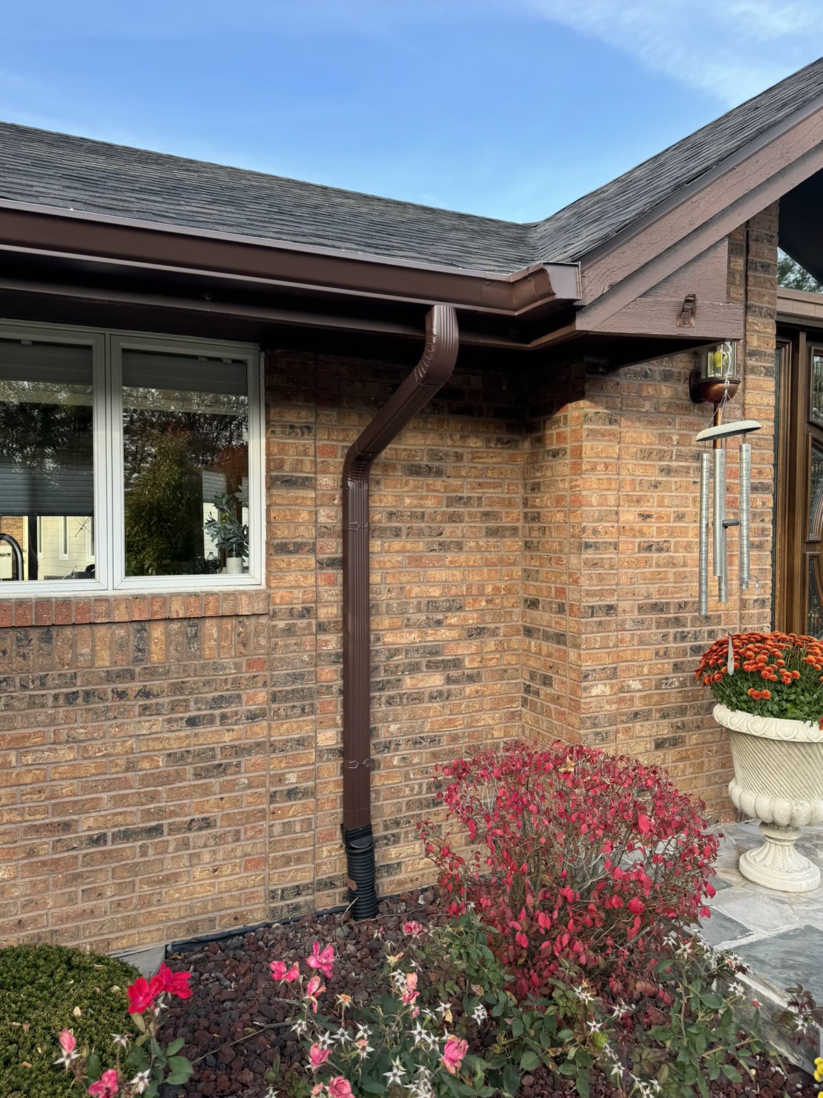 Brown seamless K-style gutter and new downspout installed on a brick ranch home, downspout running into a landscaped flower bed