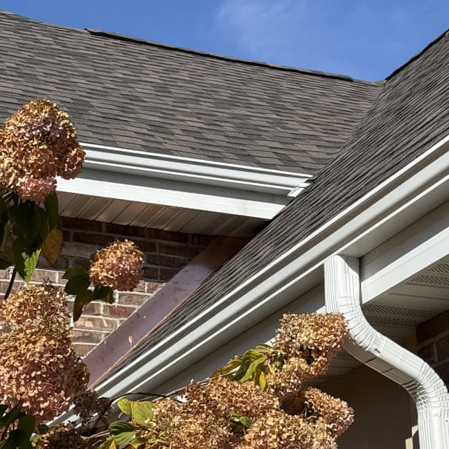 White seamless aluminum K-style gutter installed on a brown brick home with a dark asphalt shingle roof, photographed under a clear blue sky
