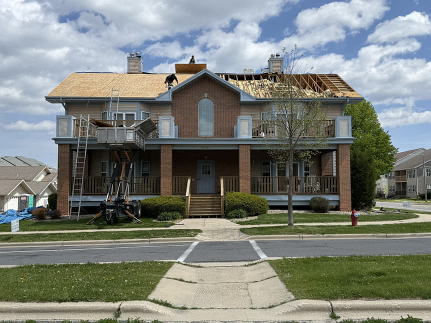 Two-story brick home mid roof replacement, debris boom in the driveway and crew working on a tear-off, Madison area Wisconsin